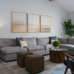 A modern living room features a large gray sectional, two brown ottomans, a round glass coffee table with a wood base, a potted plant, and the Desert Haze Set of 3 minimalist wall art above the sofa for stylish decor.
