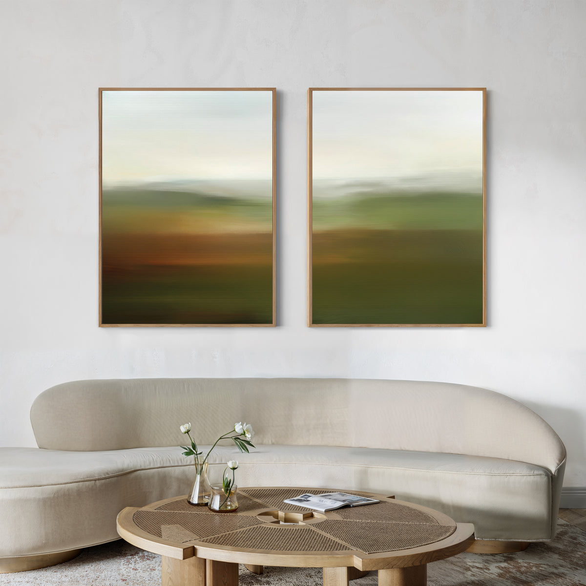 A modern living room features a curved beige sofa, a round wooden coffee table with a vase of white flowers, and the Lush Landscape Set of 2 abstract wall art prints in earthy tones displayed on a light-colored wall.