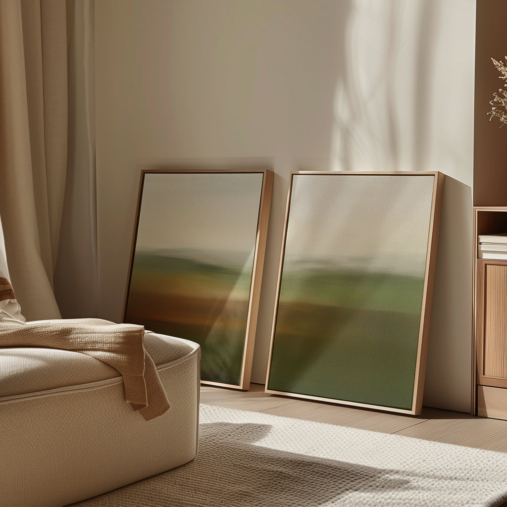 The Lush Landscape Set of 2 framed prints, featuring soft green, brown, and beige tones, rests on the floor against a light wall in a cozy sunlit room with neutral decor and a cream armchair.