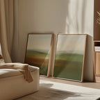 The Lush Landscape Set of 2 framed prints, featuring soft green, brown, and beige tones, rests on the floor against a light wall in a cozy sunlit room with neutral decor and a cream armchair.