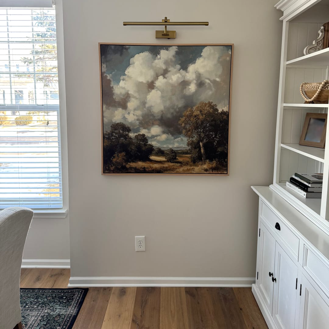 The Gathering Storm Canvas Art, featuring dramatic clouds and trees, hangs on a beige wall under a brass picture light above a white cabinet. The room also includes books, wood floors, blinds, and the corner of a patterned rug.