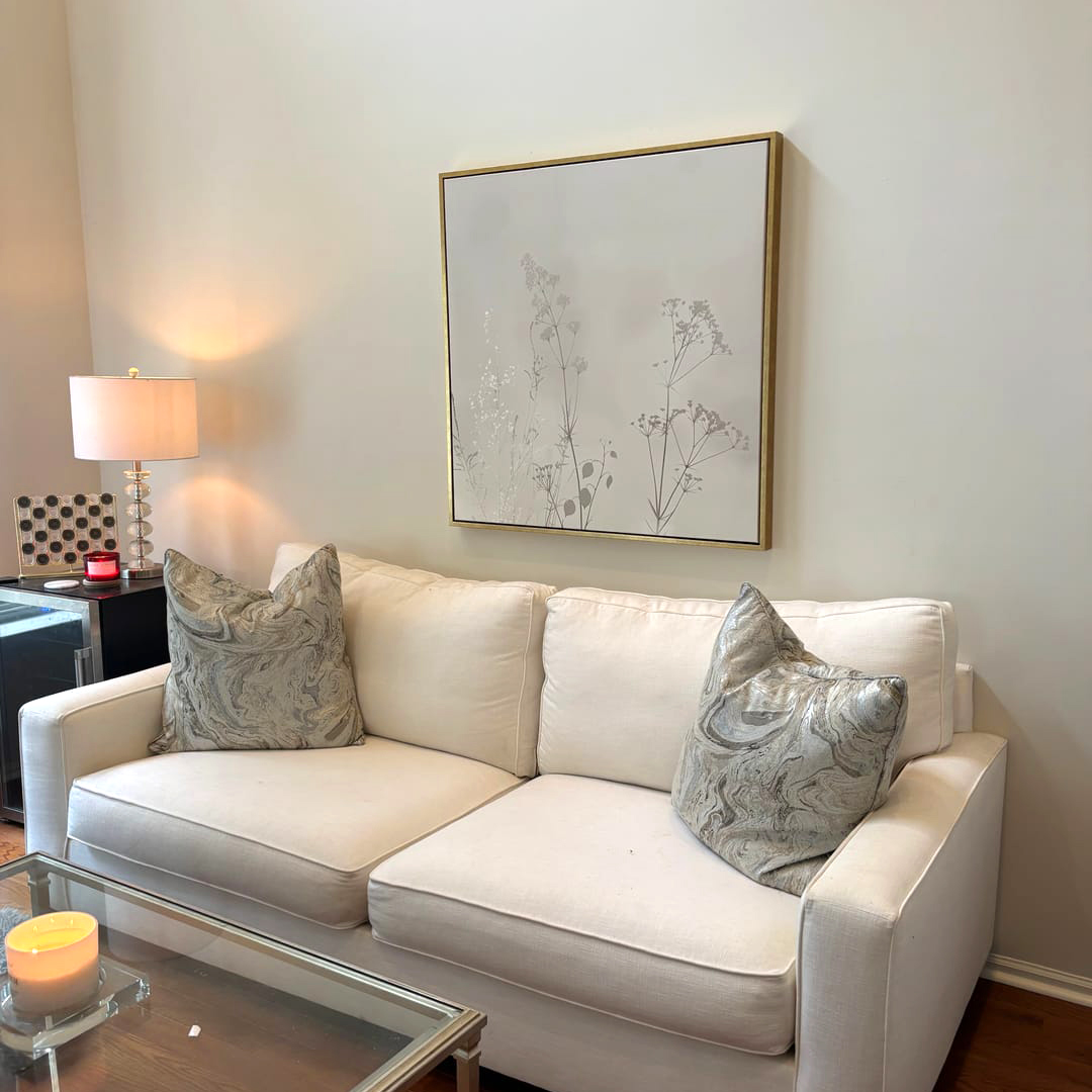 A modern living room with a cream-colored sofa, two patterned pillows, a glass coffee table with a candle, a lamp on a side table, and the Floral Symphony Abstract Canvas Art above the sofa for stylish decor.