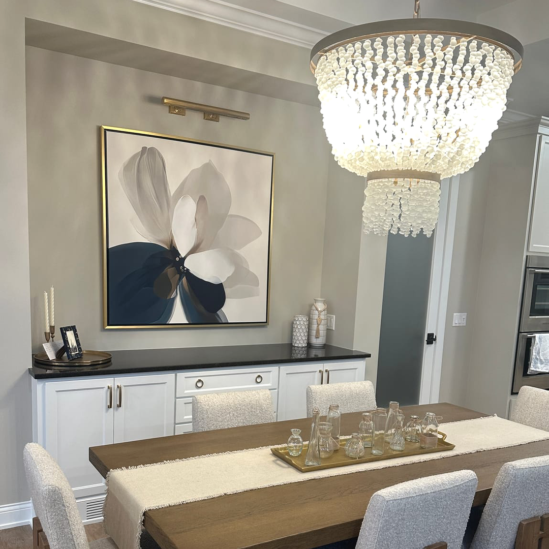 A modern dining room features a wooden table set for drinks, upholstered chairs, a beaded chandelier, serene wall art above white cabinetry with decor accents, and the Whispering Petals Canvas Art elevating the elegant ambiance.
