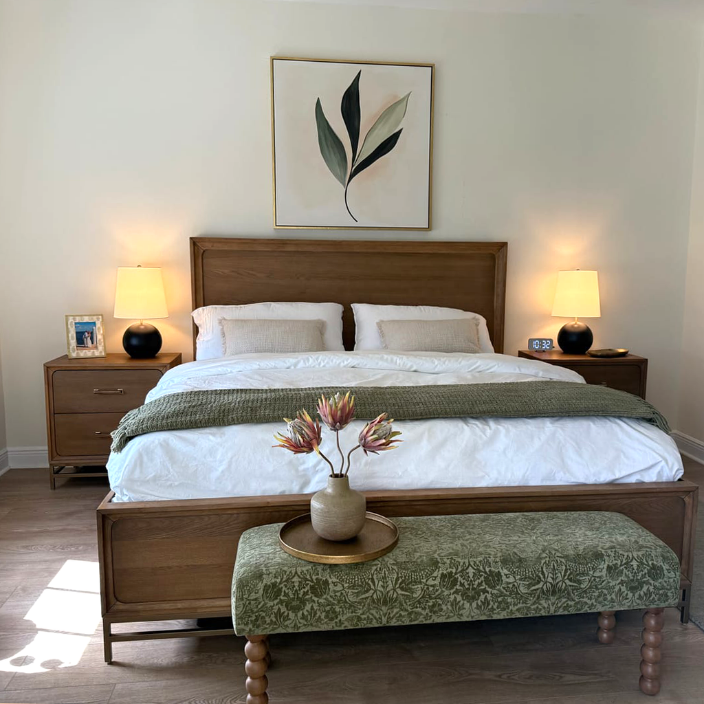 A cozy bedroom features a wooden bed with white bedding, green throw, two bedside tables with lamps, a green bench, and the Silent Leaf Canvas Art framed above the bed. A vase of flowers sits on a tray for added charm.