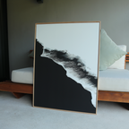 The Black Ocean Abstract Canvas Art, featuring bold black and white contrasts in an abstract wave or mountain design, is framed and leans against a cushioned wooden bench indoors.