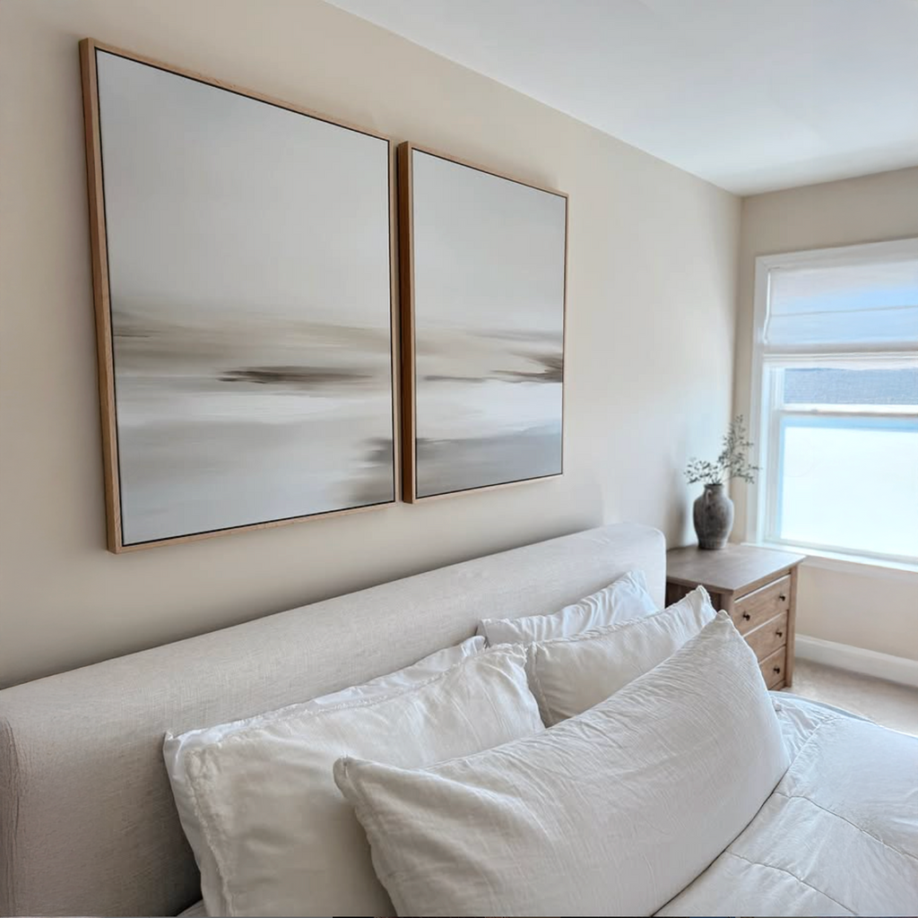 A bright, minimalist bedroom features a beige bed with white bedding, a wooden nightstand, and natural light from a window. Above the headboard hangs the Tranquil Shifts Set of 2 wall art pieces for a modern touch.