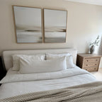A neatly made bed with white bedding sits against a beige wall, above which hangs the Tranquil Shifts Set of 2 modern wall art. A wooden nightstand with a vase of greenery stands to the right of the bed.