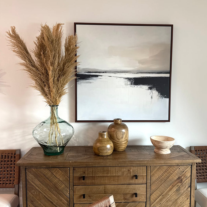 A wooden sideboard displays a glass vase with pampas grass, two wooden vases, and a bowl. Above hangs the Binary Calm Abstract Canvas Art in neutral tones, completing this modern decor scene with natural accents.