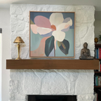 A white stone fireplace with a wooden mantel exudes modern elegance, featuring the Bold Bloom Canvas Art, a brass lamp, and a small Buddha statue. A bookshelf is partially visible on the right.