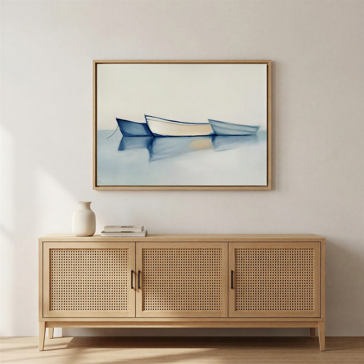 A minimalist room features a light wooden sideboard, a white vase, and two books. Above, the "Harbor Rest Canvas Art - Horizontal" depicts three blue rowboats on calm water—a tranquil maritime accent against a neutral backdrop.