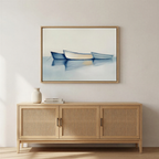 A minimalist room features a light wooden sideboard, a white vase, and two books. Above, the "Harbor Rest Canvas Art - Horizontal" depicts three blue rowboats on calm water—a tranquil maritime accent against a neutral backdrop.