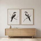 The Still Perch Set of 2, framed black-and-white bird illustrations, hangs above a light wood console with a ceramic vase, set against a neutral wall—ideal perched birds decor for a minimalist room.
