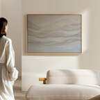 A woman in a white robe stands in a minimal, modern room, looking at the Sandy Hues Abstract Canvas Art with soft wavy lines in neutral tones, displayed above a light-colored, textured sofa.