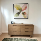 A wooden dresser with six drawers stands against a white wall. Above it hangs the Petal Hues Canvas Art, while a small framed photo and a few small items rest on the dresser.