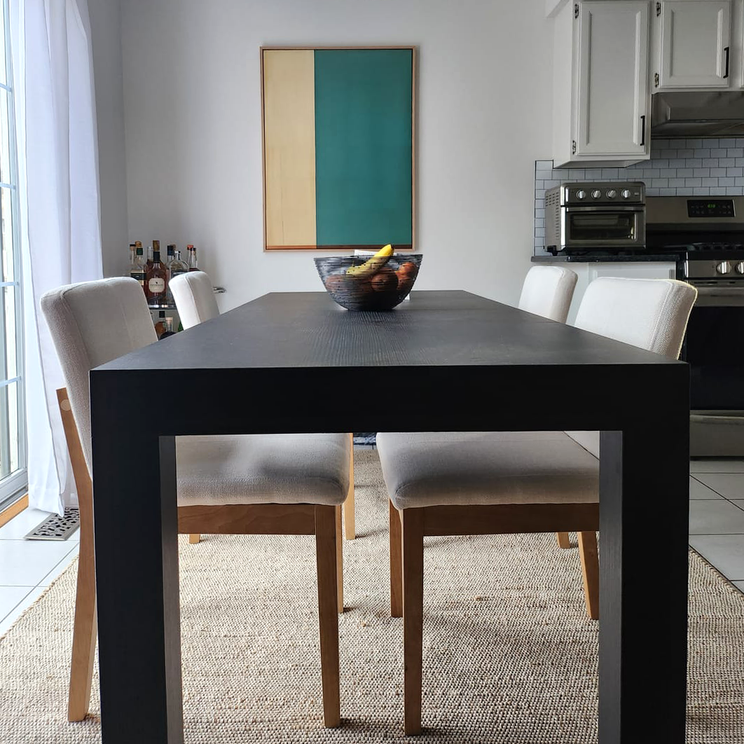 A modern dining room with a black rectangular table, four beige chairs, a bowl of fruit in the center, and Color Division Abstract Canvas Art hanging on a white wall in the background.