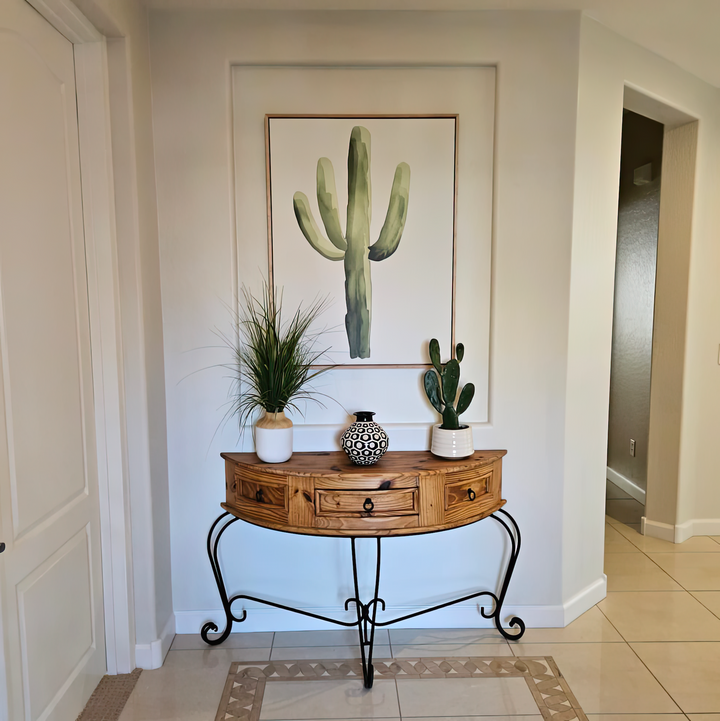 A wooden console table with black metal legs sits against the wall, topped with two potted plants and a decorative vase. Above it hangs the Desert Cactus Canvas Art—a perfect botanical accent for minimalist decor.