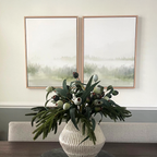 A white textured vase with green foliage sits on a dining table, while the Misty Forest Set of 2 framed paintings depicting misty green forests hang on a light wall above a gray chair—perfect for serene, natural accents.