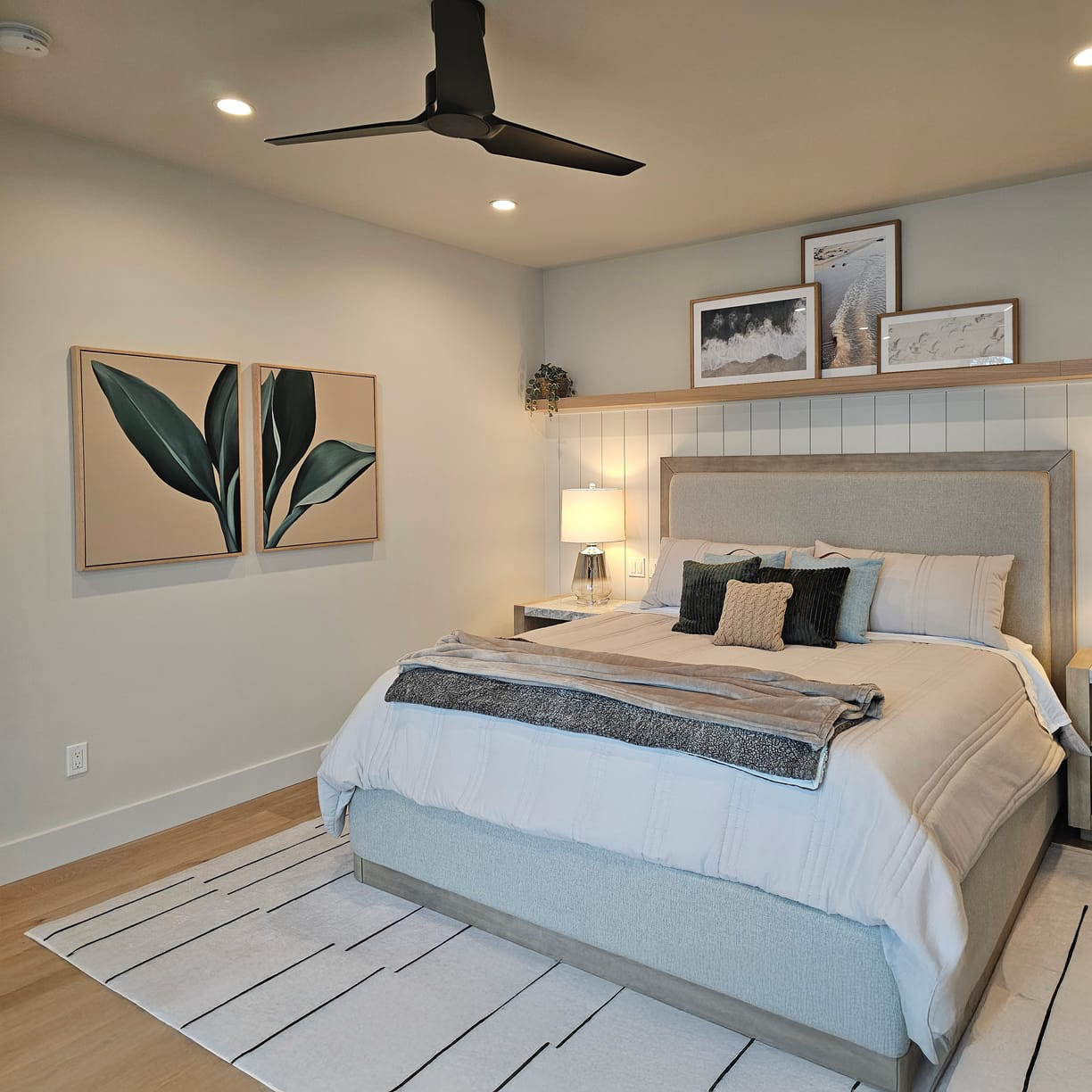 A modern bedroom with a neatly made bed, neutral bedding, a gray upholstered headboard, two framed leaf prints on one wall, and a shelf with framed art and a potted plant above the bed, under ceiling spotlights and a black fan.