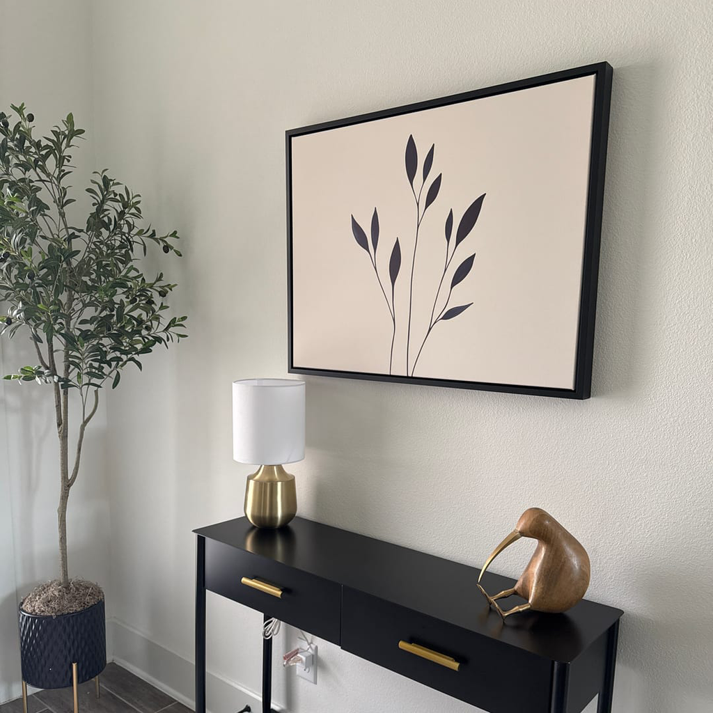 A minimalist entryway features a black console table with brass accents, a gold and white lamp, a wooden bird sculpture, a potted plant, and modern wall decor with the Navy Stem Canvas Art on a light-colored wall.