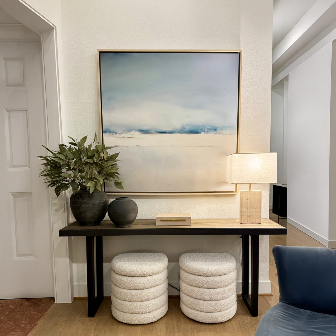 A modern entryway featuring a dark wood console, two round white ottomans, a leafy plant in a black vase, wicker lamp, and tranquil home decor highlighted by the Misty Morning Abstract Canvas Art in soft blue and white tones.
