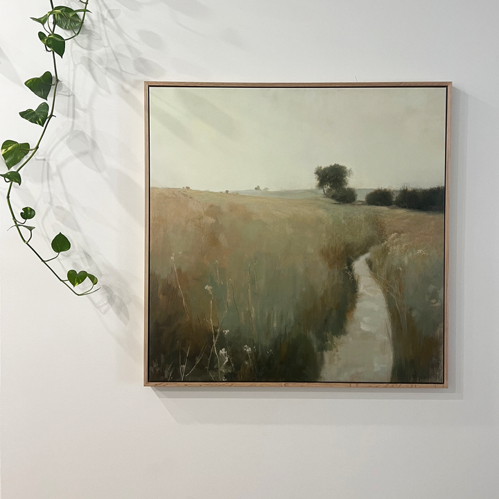 Path Through the Meadow Canvas Art features a grassy field with a winding path and distant trees, framed and hung on a white wall, accented by a leafy green vine trailing from the top left—ideal décor for nature lovers.