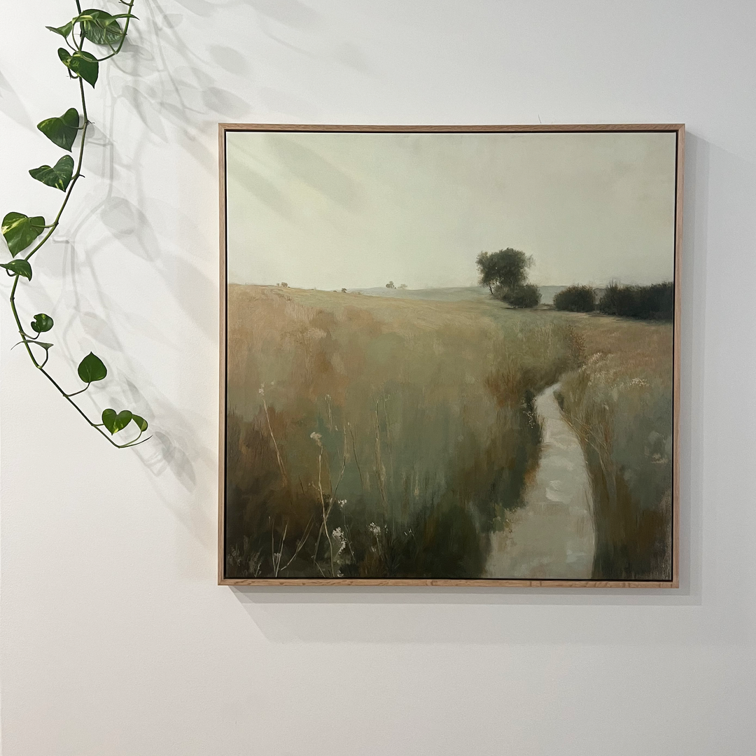 The Path Through the Meadow Canvas Art features a grassy field with a winding path, framed and hung on a white wall. A leafy vine drapes along the top edge, casting gentle shadows—ideal rustic wall decor for nature lovers.