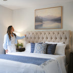 A woman stands by a bed with blue and white bedding, touching the tufted beige headboard. Above the bed hangs the Smoky Peaks Canvas Art, depicting mountains and a lake, giving the bright room a cozy, vintage feel.