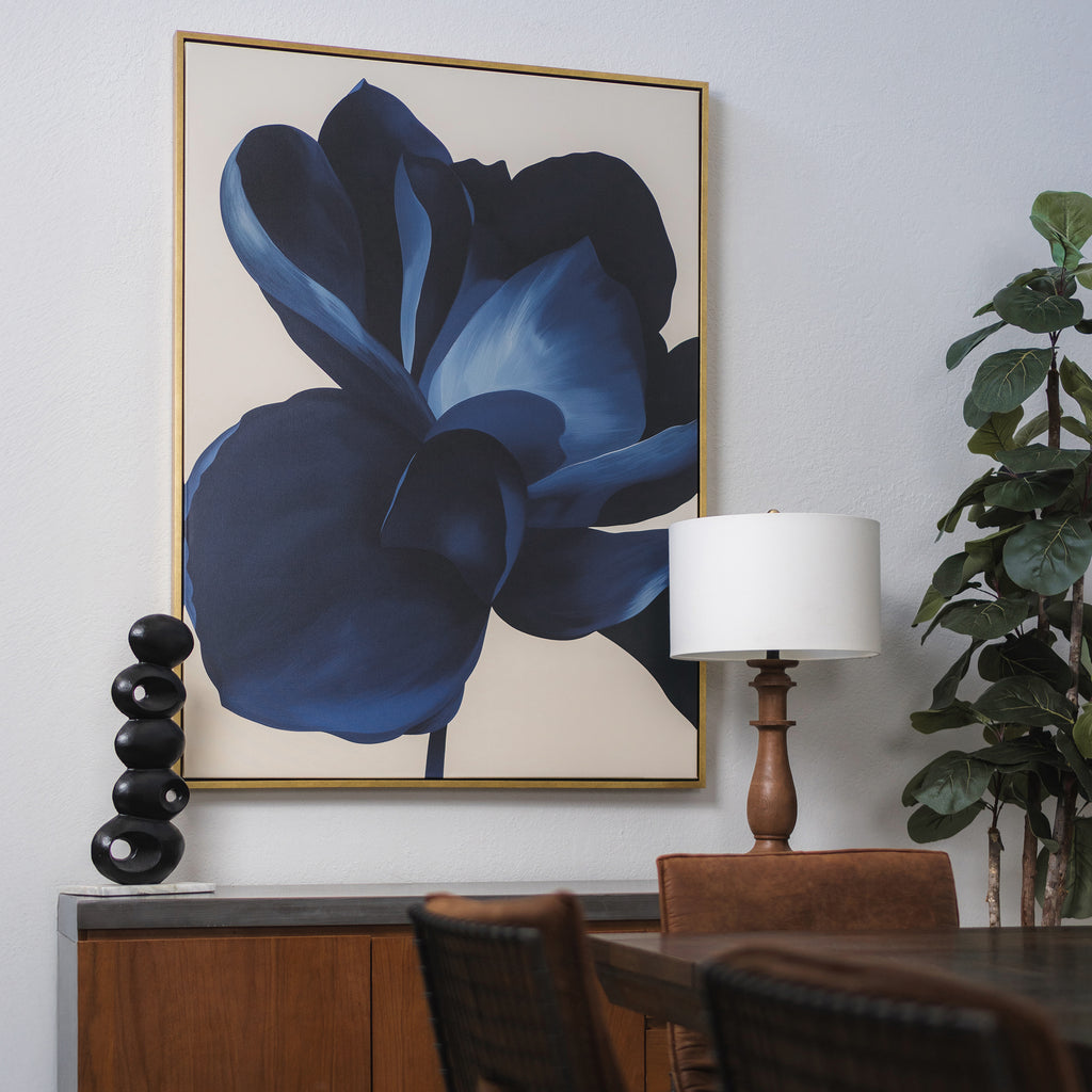 The Midnight Bloom Abstract Canvas Art, featuring a large blue flower, hangs above a wooden sideboard on a light gray wall. Modern décor accents like a black sculpture, table lamp, and green plant complete the stylish dining area.