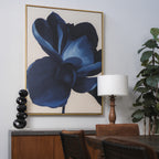 The Midnight Bloom Abstract Canvas Art, featuring a large blue flower, hangs above a wooden sideboard on a light gray wall. Modern décor accents like a black sculpture, table lamp, and green plant complete the stylish dining area.