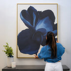 A woman with long dark hair hangs the Midnight Bloom Abstract Canvas Art—a large blue floral piece—on a light wall above a table decorated with minimalist greenery and a lit candle.