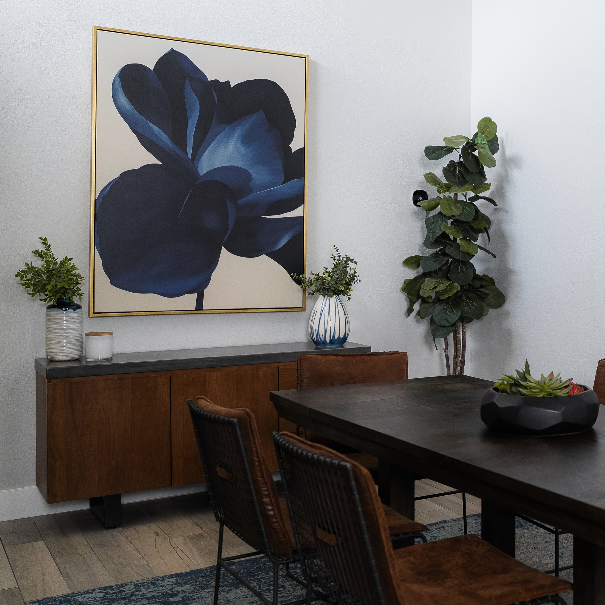 A modern dining room featuring a dark wood table, wicker chairs, sideboard, potted plants, and the Midnight Bloom Abstract Canvas Art—a striking blue floral painting that adds contemporary flair as wall decor.