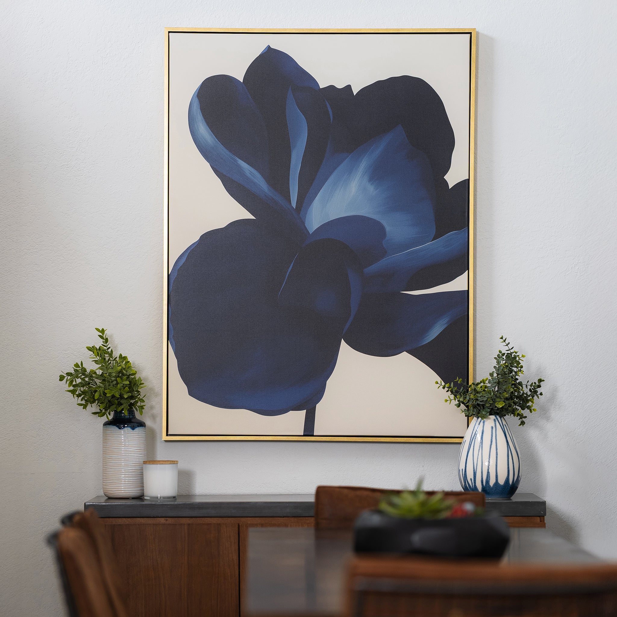 A framed Midnight Bloom Abstract Canvas Art featuring a large blue flower hangs on a white wall above a wooden table adorned with a small plant, two candles, and a white vase with green foliage, enhancing the modern wall decor.