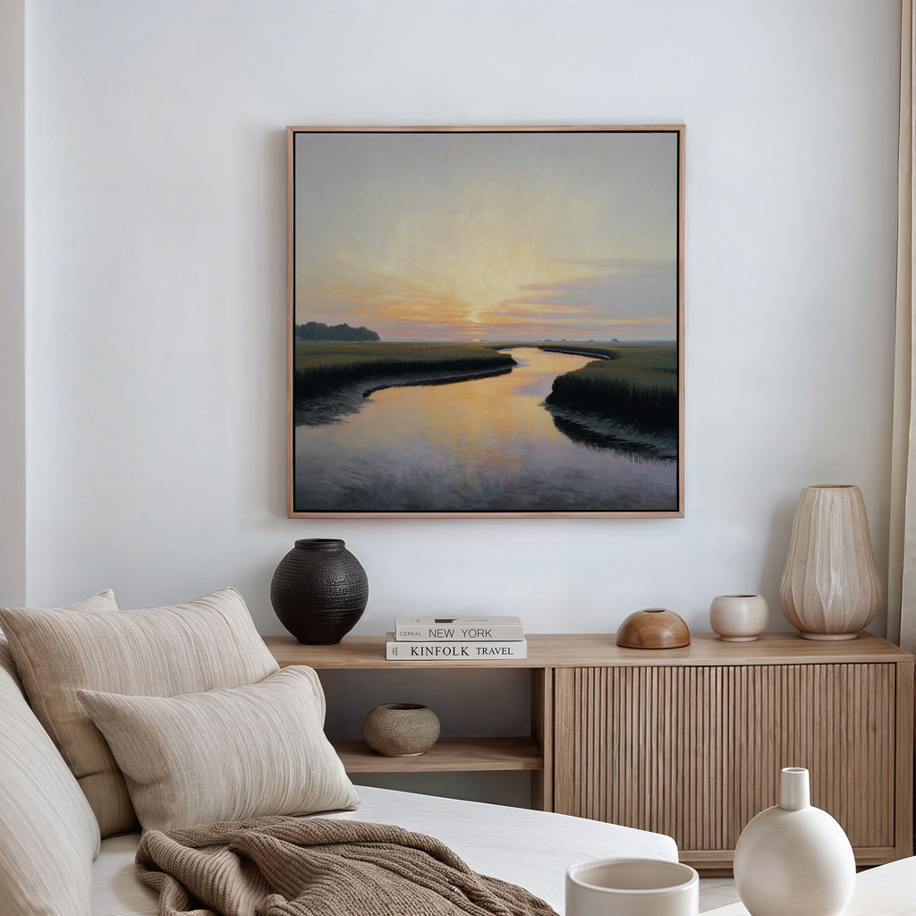 A serene living room with neutral decor showcases the Tidal Whispers Canvas Art - Square, depicting a sunset over a river above a wooden console with books and vases. A beige sofa with striped cushions is in the foreground.