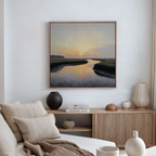 A serene living room with neutral decor showcases the Tidal Whispers Canvas Art - Square, depicting a sunset over a river above a wooden console with books and vases. A beige sofa with striped cushions is in the foreground.