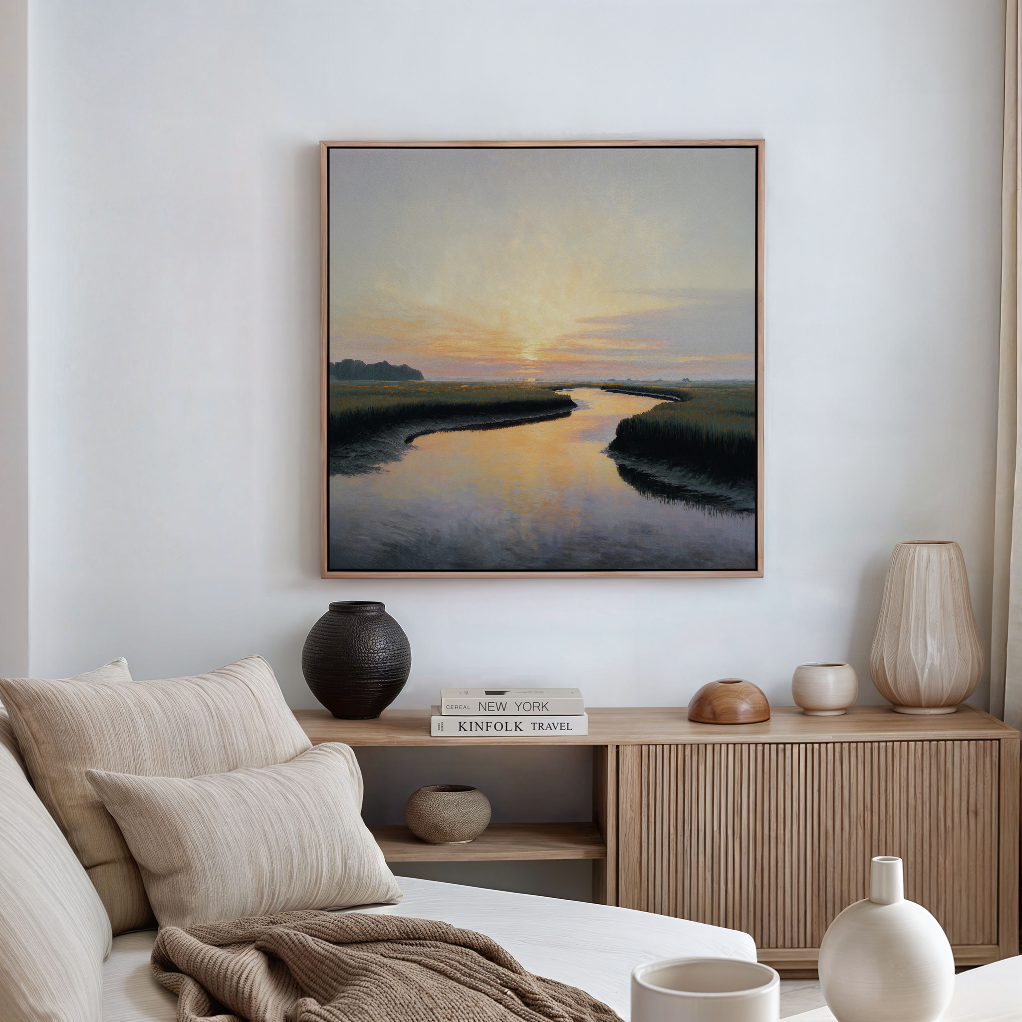 A serene living room with neutral decor showcases the Tidal Whispers Canvas Art - Square, depicting a sunset over a river above a wooden console with books and vases. A beige sofa with striped cushions is in the foreground.