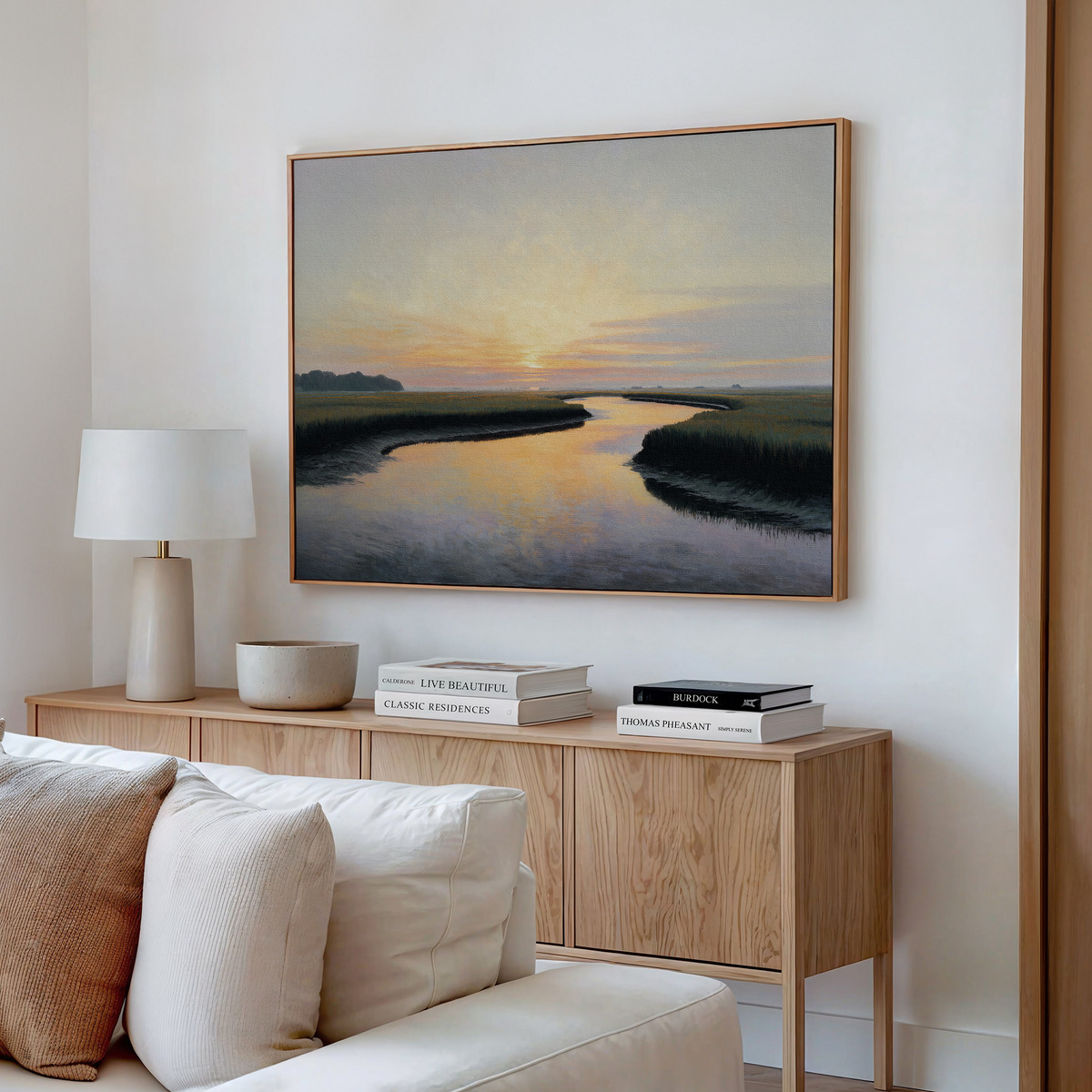 A modern living room features a pale sofa, wooden sideboard with books and ceramics, white lamp, and the Tidal Whispers Canvas Art - Horizontal depicting a serene river at sunset displayed on a white wall.