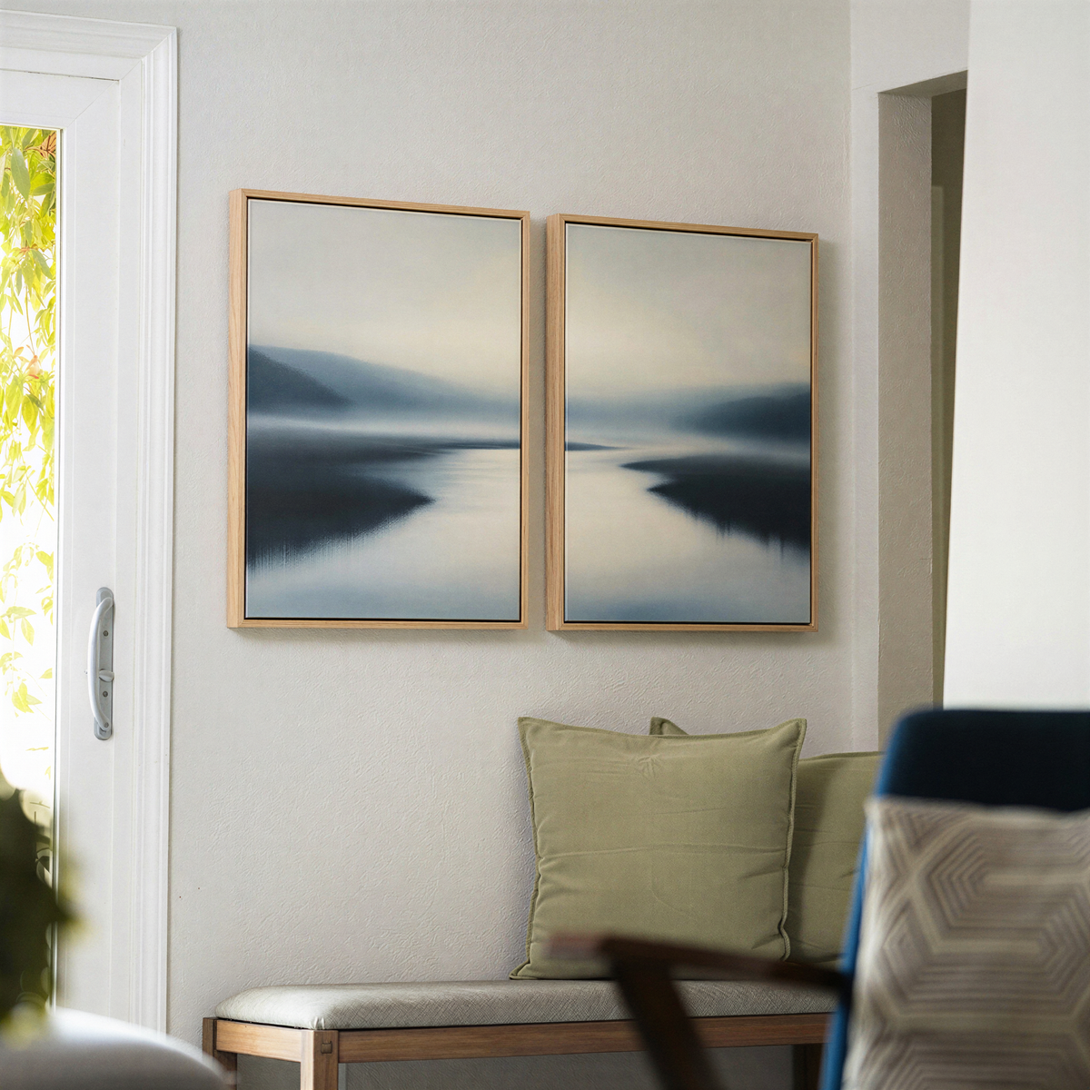 The "Peaceful Waters Set of 2" framed landscape wall art paintings, featuring a serene misty river scene, are displayed on a light-colored wall above a bench with green pillows in a softly lit, modern living space, creating an atmosphere of tranquil decor.