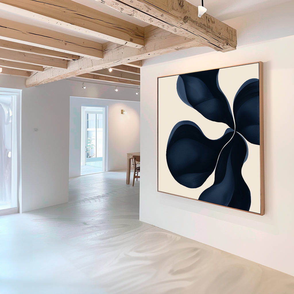 A modern, minimalist room with exposed wooden beams and white walls features Blossom Blue Abstract Canvas Art—a large print with a bold, dark blue abstract design. Natural light enhances this striking minimalist wall art.