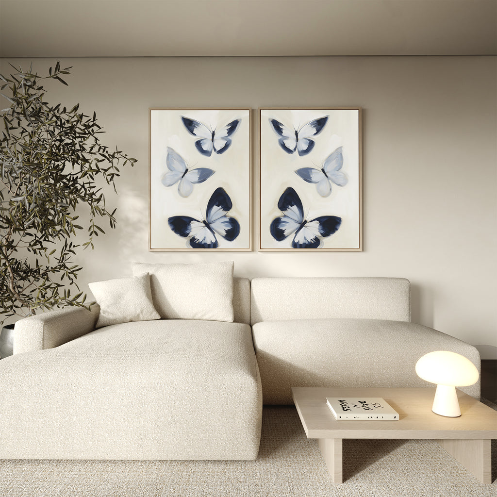 A modern living room featuring a cream sectional sofa, wooden side table with lamp and book, tall potted plant, and the chic Midnight Wings Set of 2 butterfly wall art decor on a neutral wall.