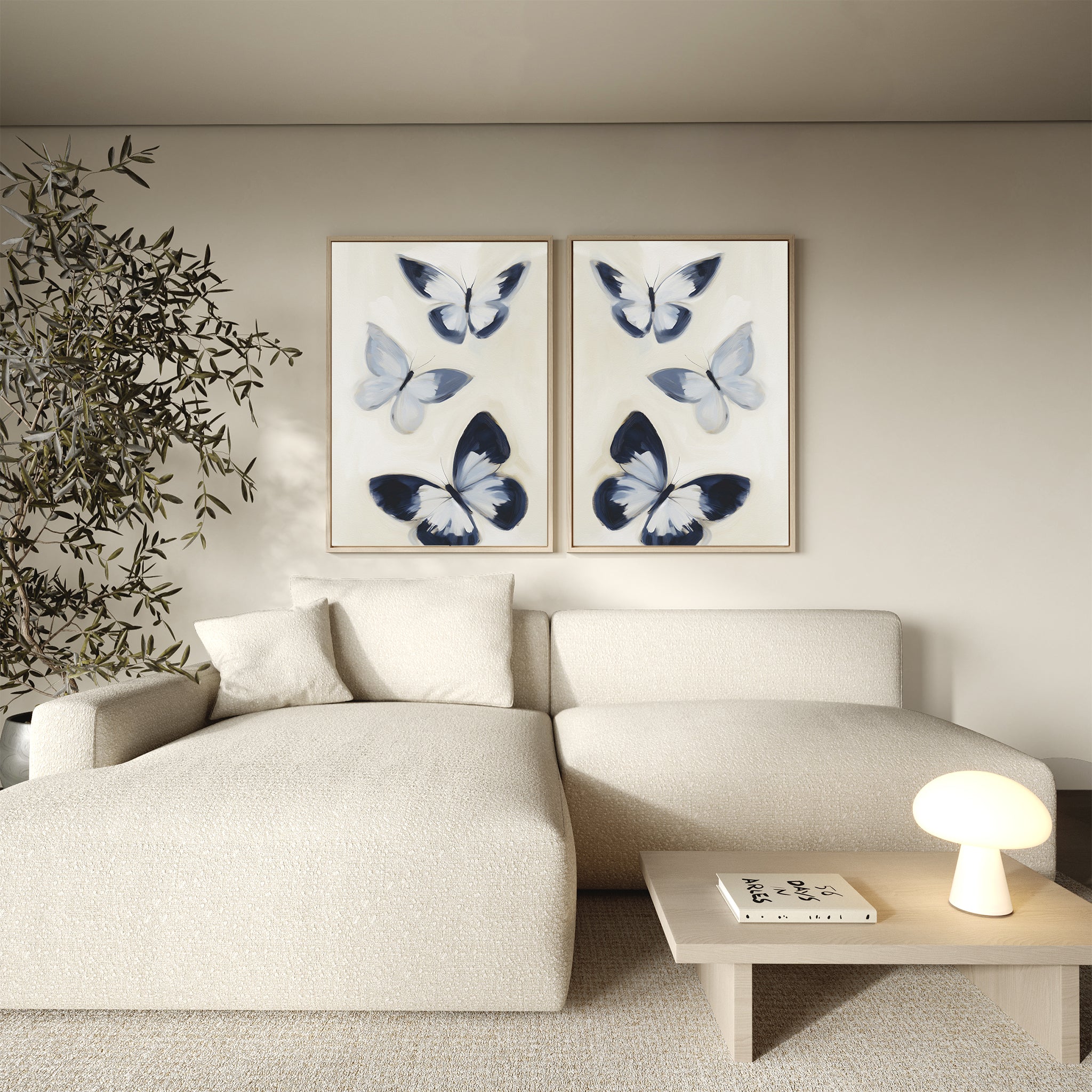 A modern living room featuring a cream sectional sofa, wooden side table with lamp and book, tall potted plant, and the chic Midnight Wings Set of 2 butterfly wall art decor on a neutral wall.