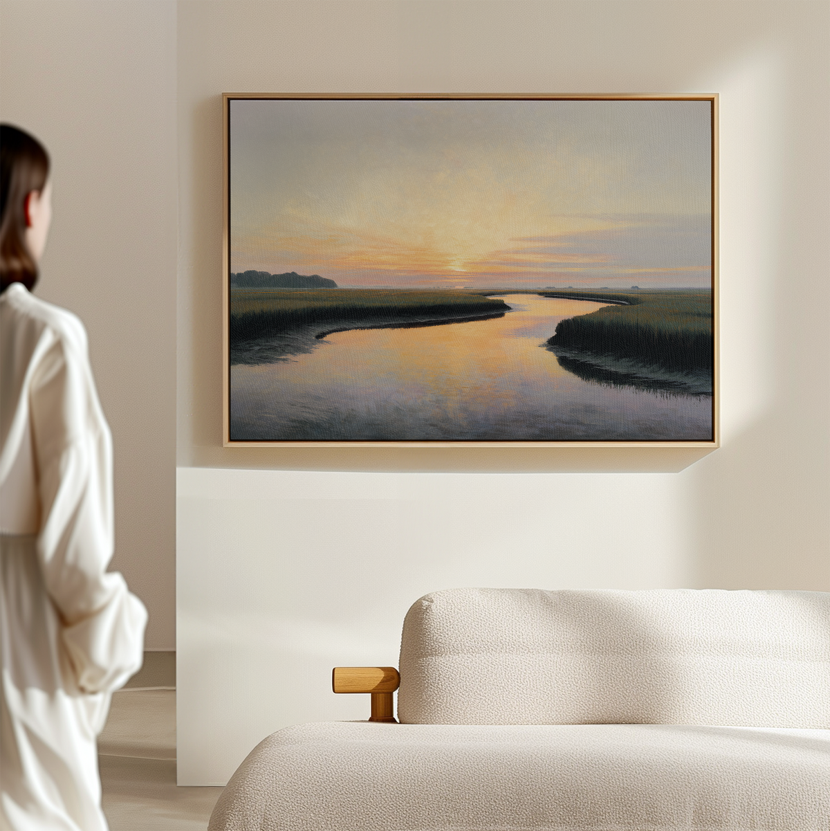 A person admires the Tidal Whispers Canvas Art - Horizontal, featuring a sunset river scene in a minimalist room. Soft sunlight reflects on water, while a light-colored sofa adds to the serene home decor.