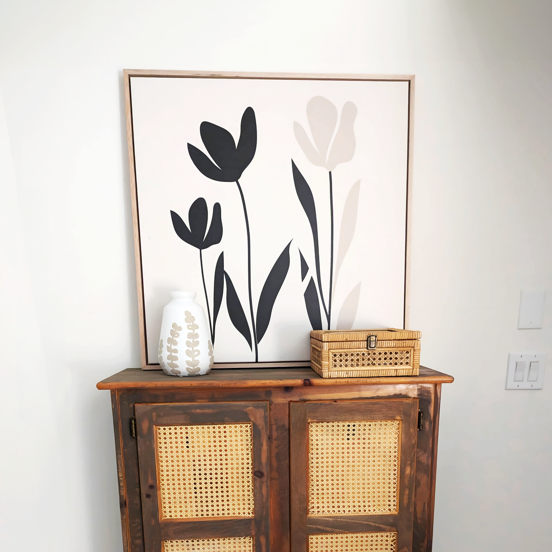A wooden cabinet with woven cane doors displays a white leaf-patterned vase and a small wicker box. Above hangs the Floral Simplicity Canvas Art, featuring three abstract black and beige flowers for a vintage-inspired touch.