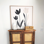 A wooden cabinet with woven cane doors displays a white leaf-patterned vase and a small wicker box. Above hangs the Floral Simplicity Canvas Art, featuring three abstract black and beige flowers for a vintage-inspired touch.