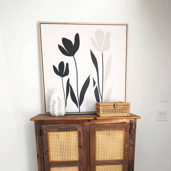 A wooden cabinet with woven cane doors displays a white leaf-patterned vase and a small wicker box. Above hangs the Floral Simplicity Canvas Art, featuring three abstract black and beige flowers for a vintage-inspired touch.