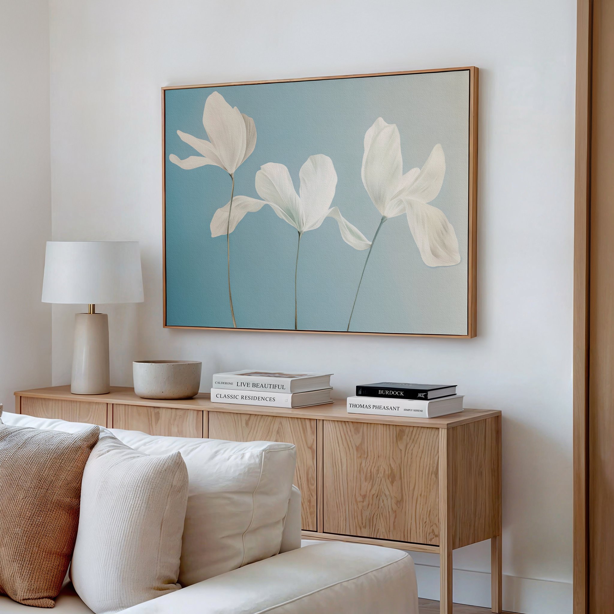 A modern living room corner features a wooden sideboard, white table lamp, stacked books, bowl, and the Stellar Flora Abstract Canvas Art showcasing white flowers on blue. A cream sofa completes the stylish decor.