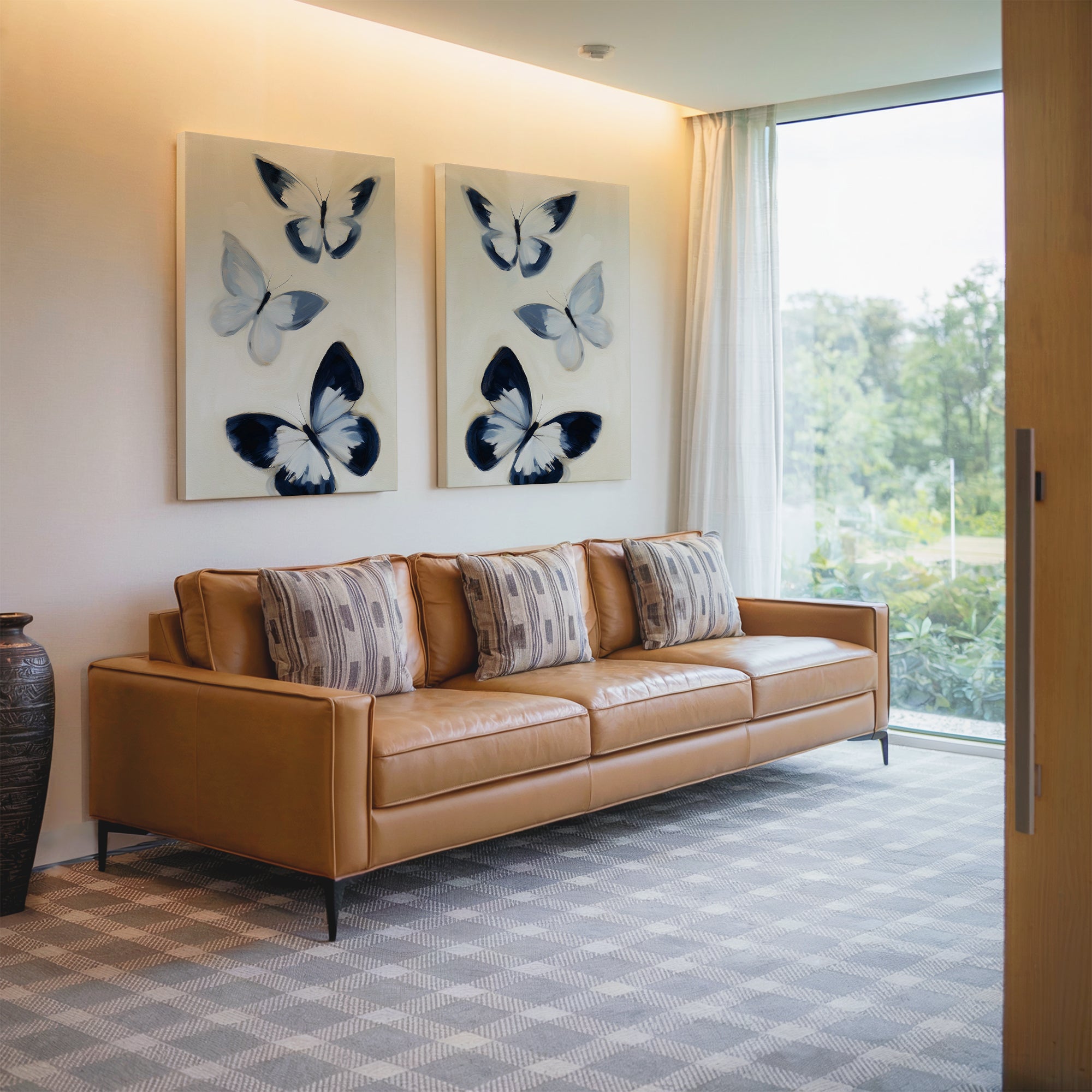 A modern living room with a tan leather sofa, four patterned pillows, Midnight Wings Set of 2 as butterfly wall art, a large window with sheer curtains, patterned carpet, and a decorative vase in the corner.