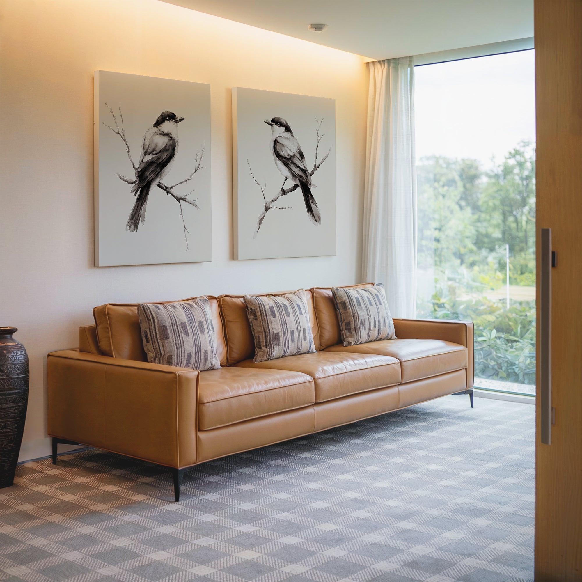 A modern living room with a tan leather sofa, patterned cushions, Still Perch Set of 2 bird art prints on the wall, a decorative vase, and large windows overlooking Nordic Park’s lush greenery.