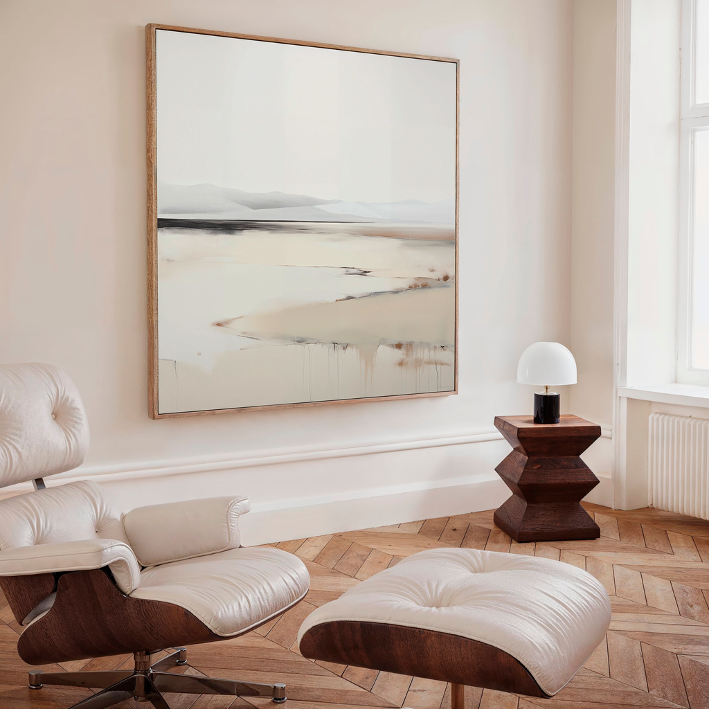 A stylish room with a cream lounge chair and ottoman, wooden herringbone floor, geometric side table with modern lamp, and Quiet Valley Abstract Canvas Art brings sophistication to the living room decor.