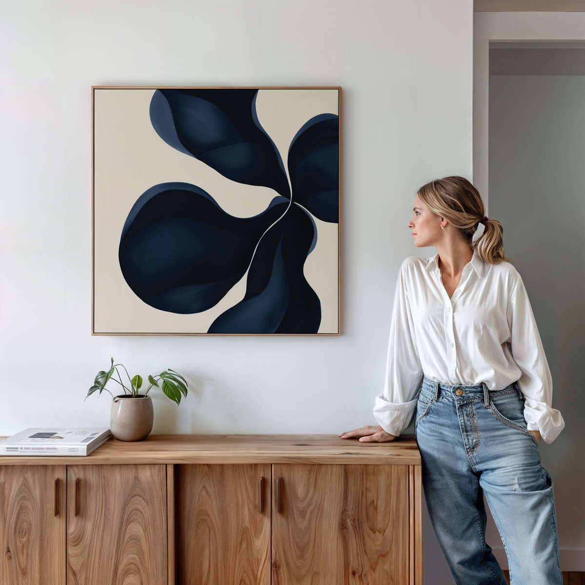 A woman in a white blouse and jeans leans on a wooden cabinet, looking at the Blossom Blue Abstract Canvas Art featuring dark blue shapes on a light background. A potted plant and books are arranged on the cabinet.