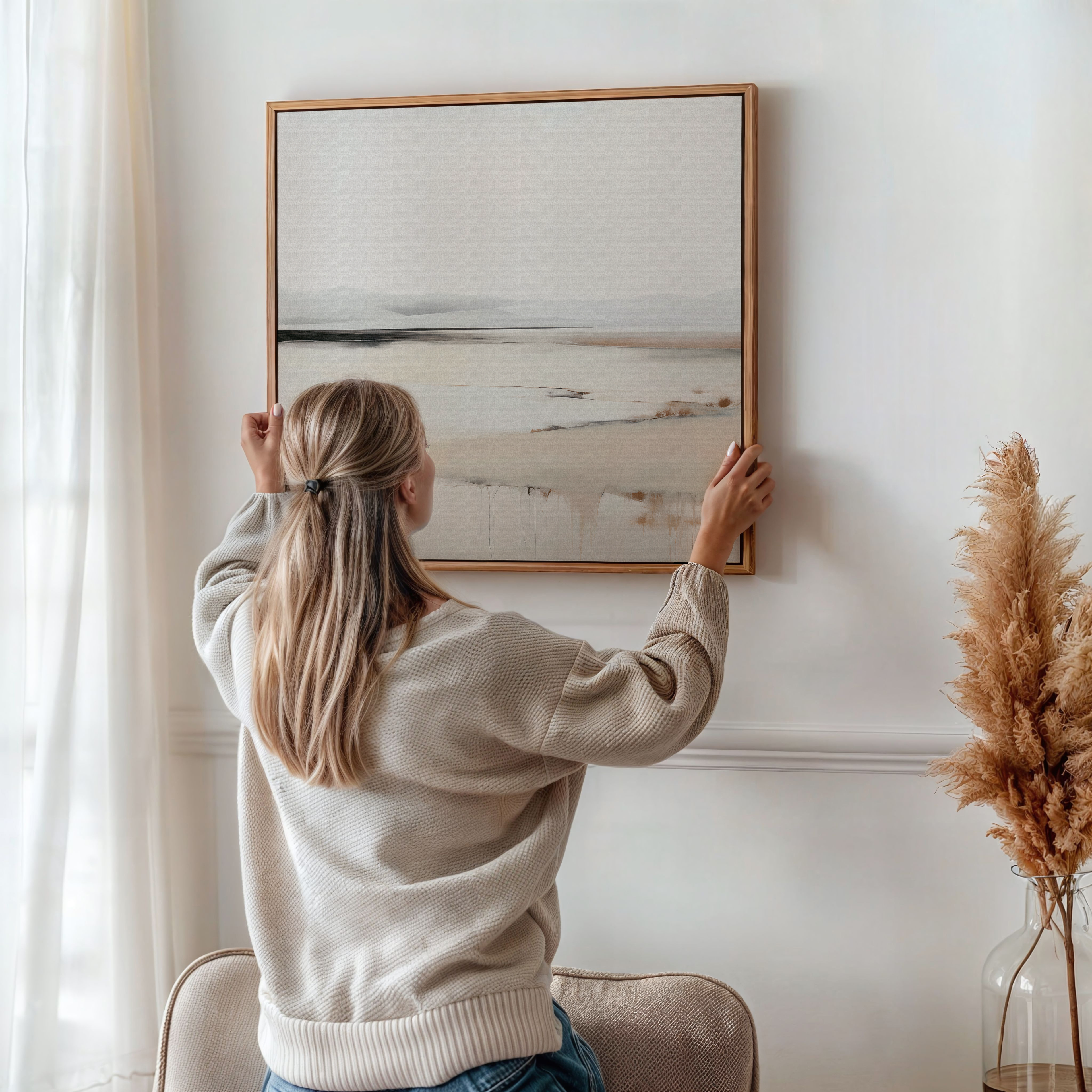 A woman with long blonde hair hangs Quiet Valley Abstract Canvas Art, perfect minimalist wall decor, on a white wall in a bright room with pampas grass and neutral accents.